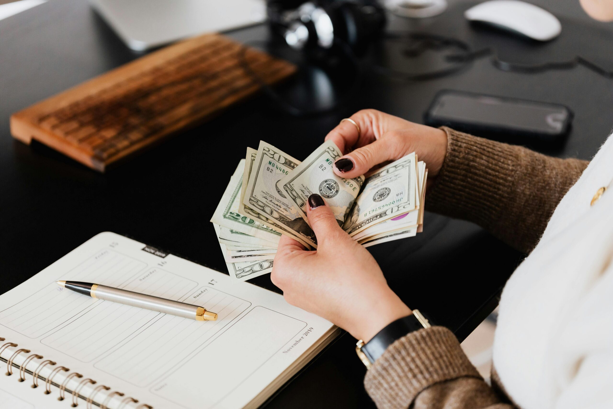 Unrecognizable elegant female in sweater counting dollar bills while sitting at wooden table with planner and pen